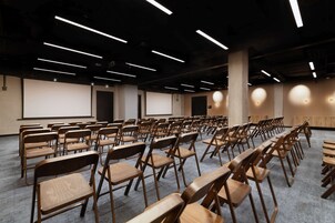 Konferenssitila, jossa on riveissä puutuoleja ja -pöytiä, projektorikangas ja modernit valaisimet.