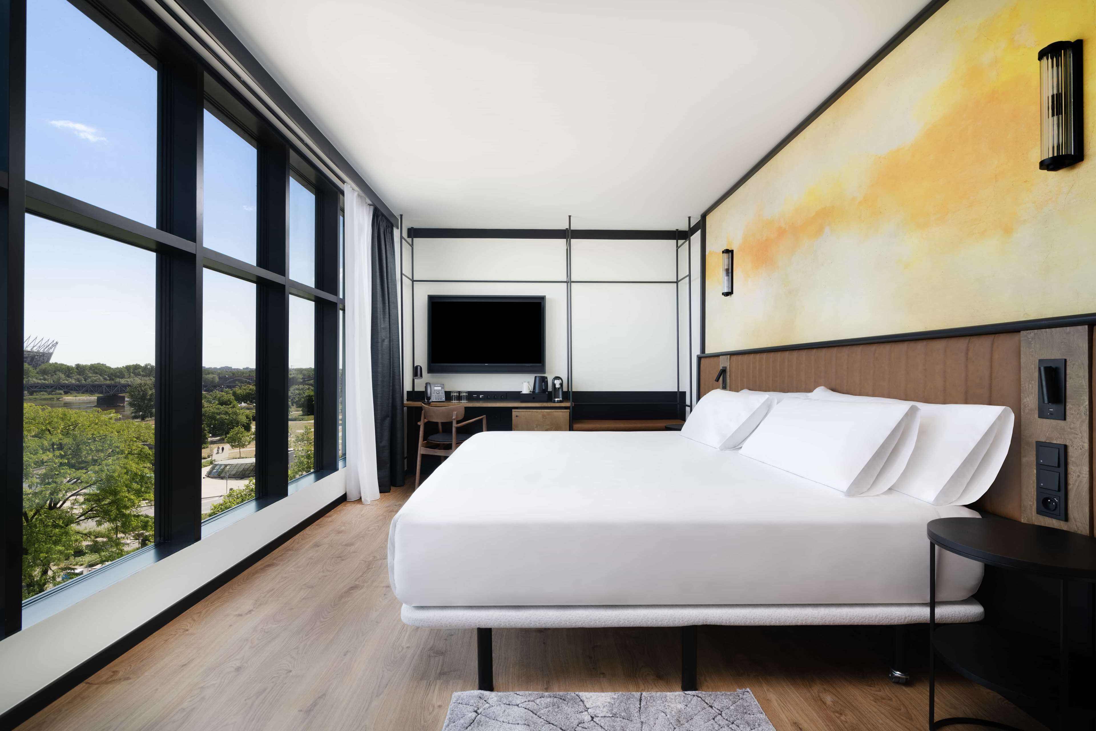 A modern hotel room with a large bed, a desk with a flat-screen TV, and a view of the outdoors through large windows.