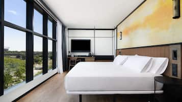 A modern hotel room with a large bed, a desk with a flat-screen TV, and a view of the outdoors through large windows.