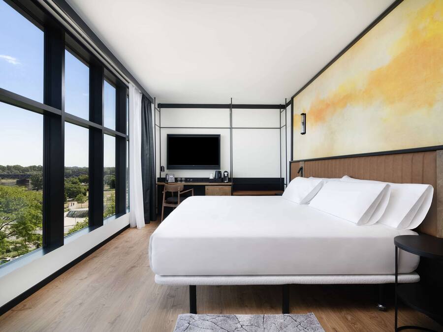A modern hotel room with a large bed, a desk with a flat-screen TV, and a view of the outdoors through large windows.