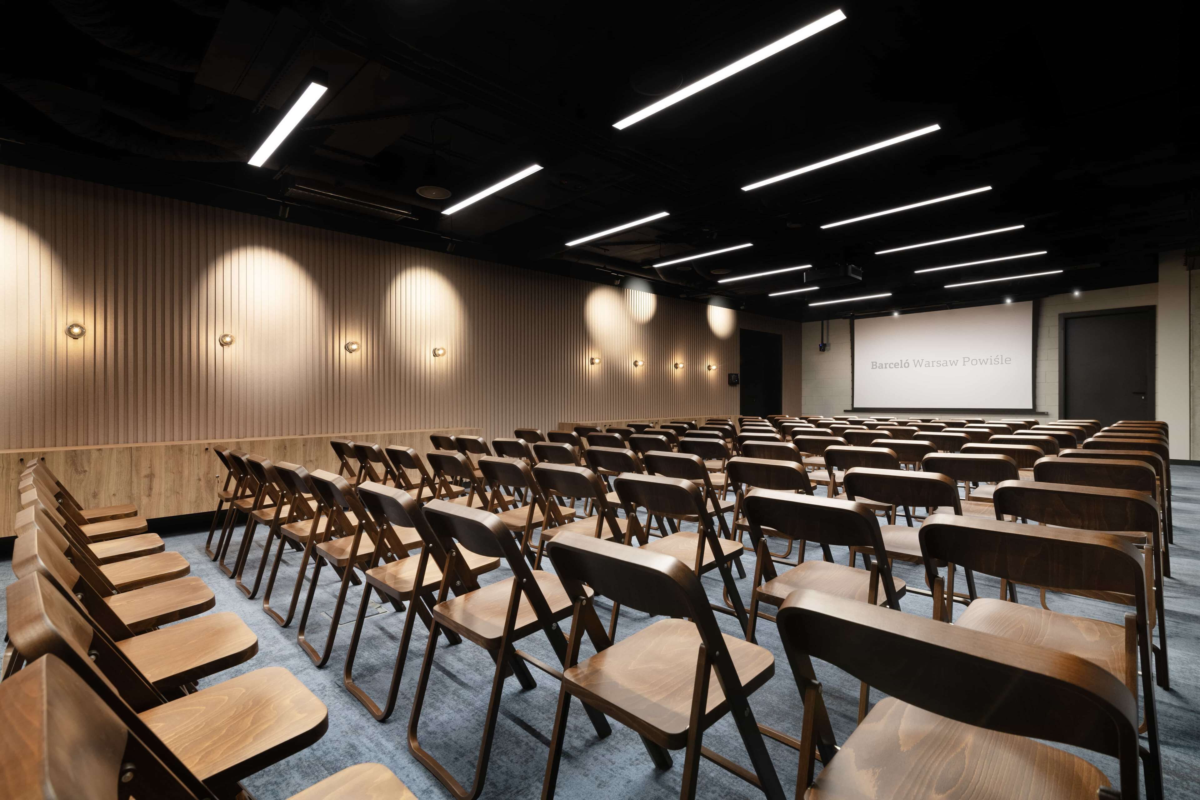 A conference room with rows of wooden chairs and a projector screen displaying "Barceló Warsaw Powiśle".