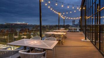 An outdoor terrace with tables and chairs, string lights, and a view of a stadium.