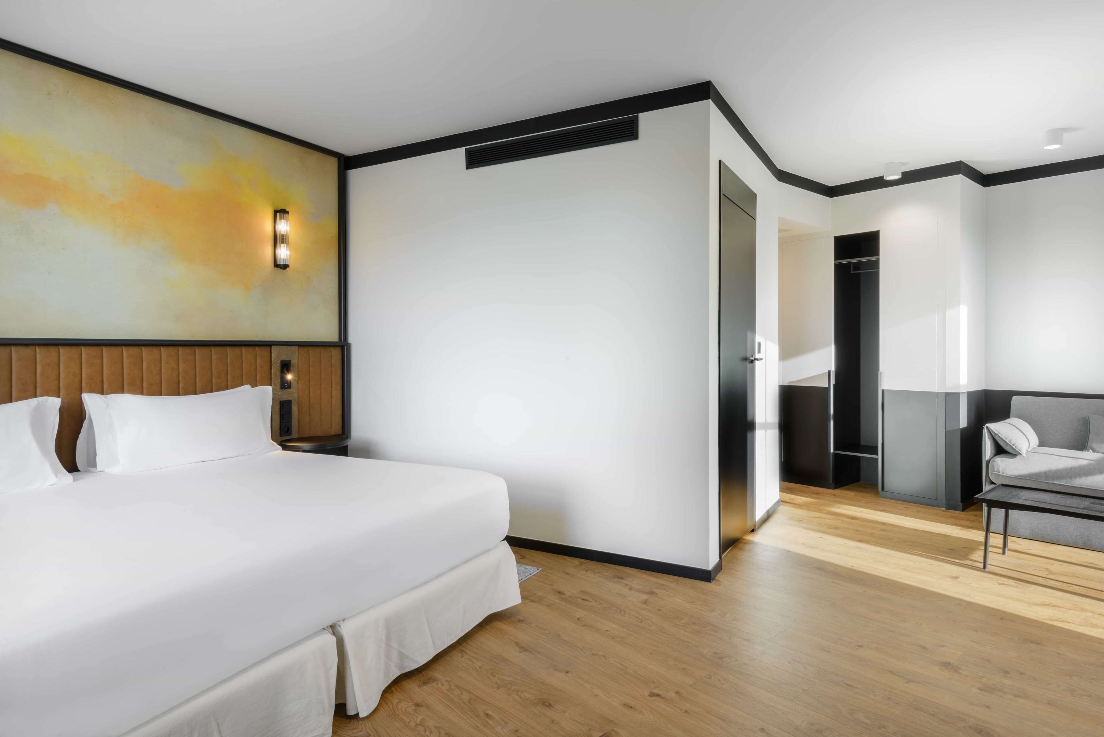 Una camera d'albergo moderna con un letto grande, una testiera in legno, una lampada a parete e un divano grigio.
