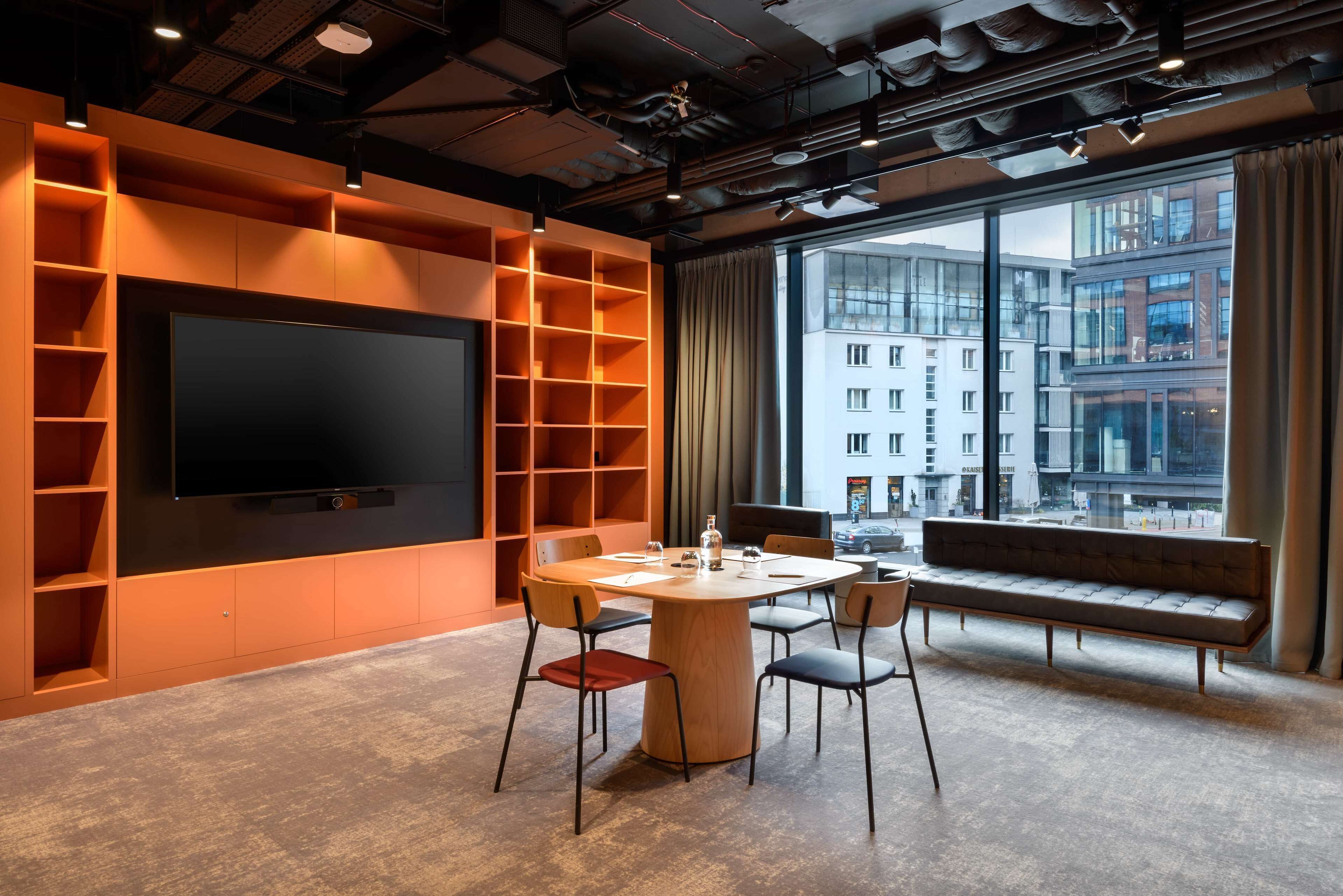 Un interior moderno con un televisor de pantalla plana grande, una mesa redonda de madera con sillas, una estantería empotrada color naranja y un ventanal con vistas a la ciudad.