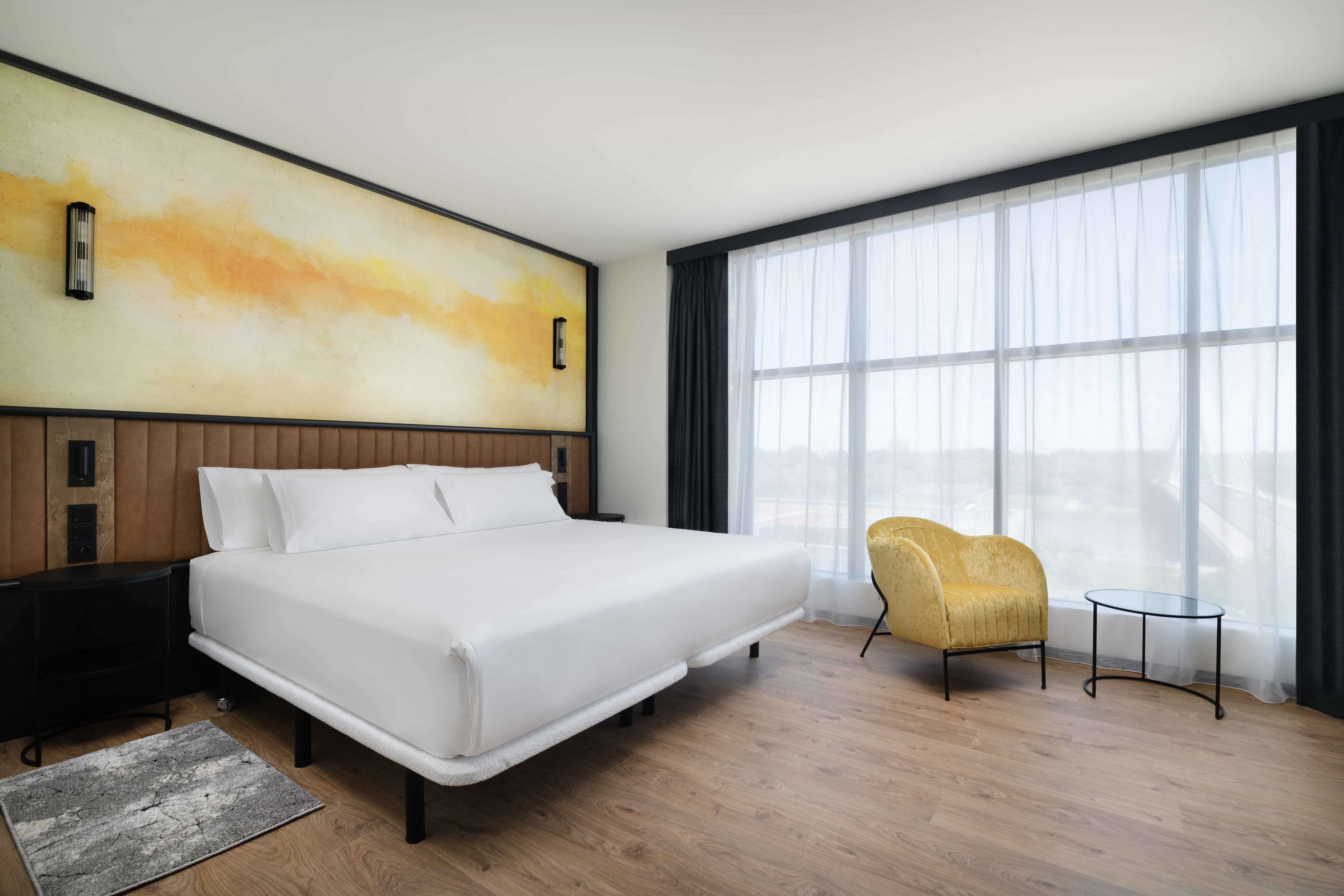 A modern hotel room with a large bed, a small round table, a yellow armchair, and a large window with sheer curtains.