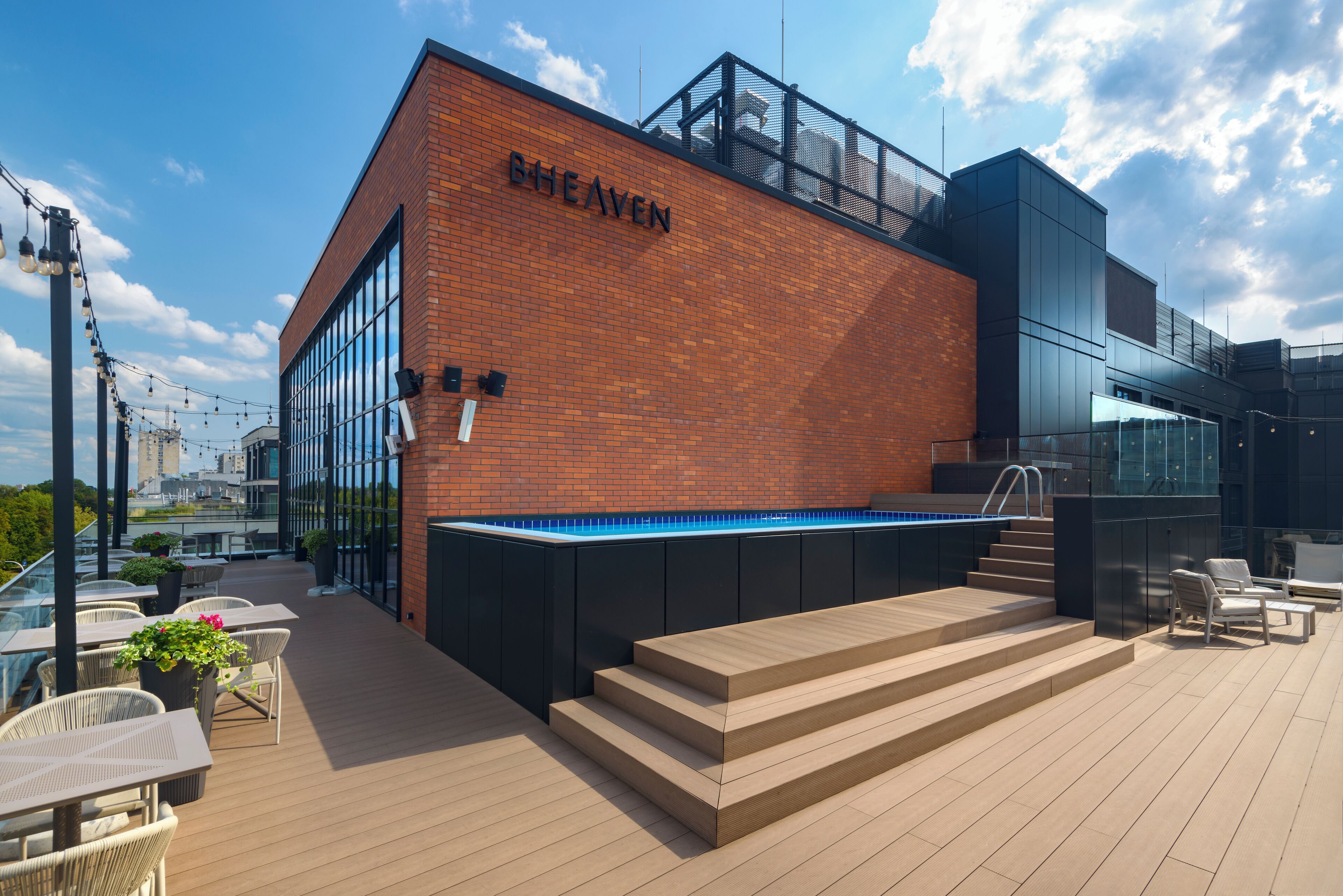 A rooftop pool area with a brick building labeled "BHEAVEN," outdoor seating, and a clear sky.