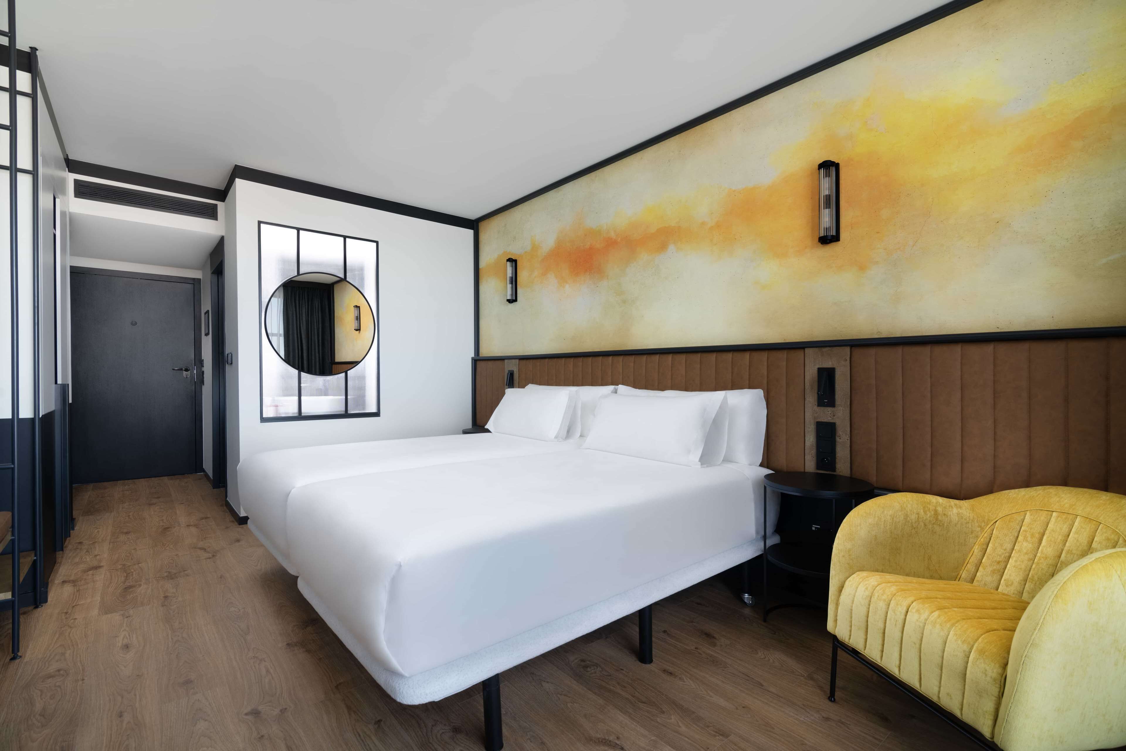 A modern hotel room with a large bed, a round mirror, a yellow armchair, and a wooden headboard.