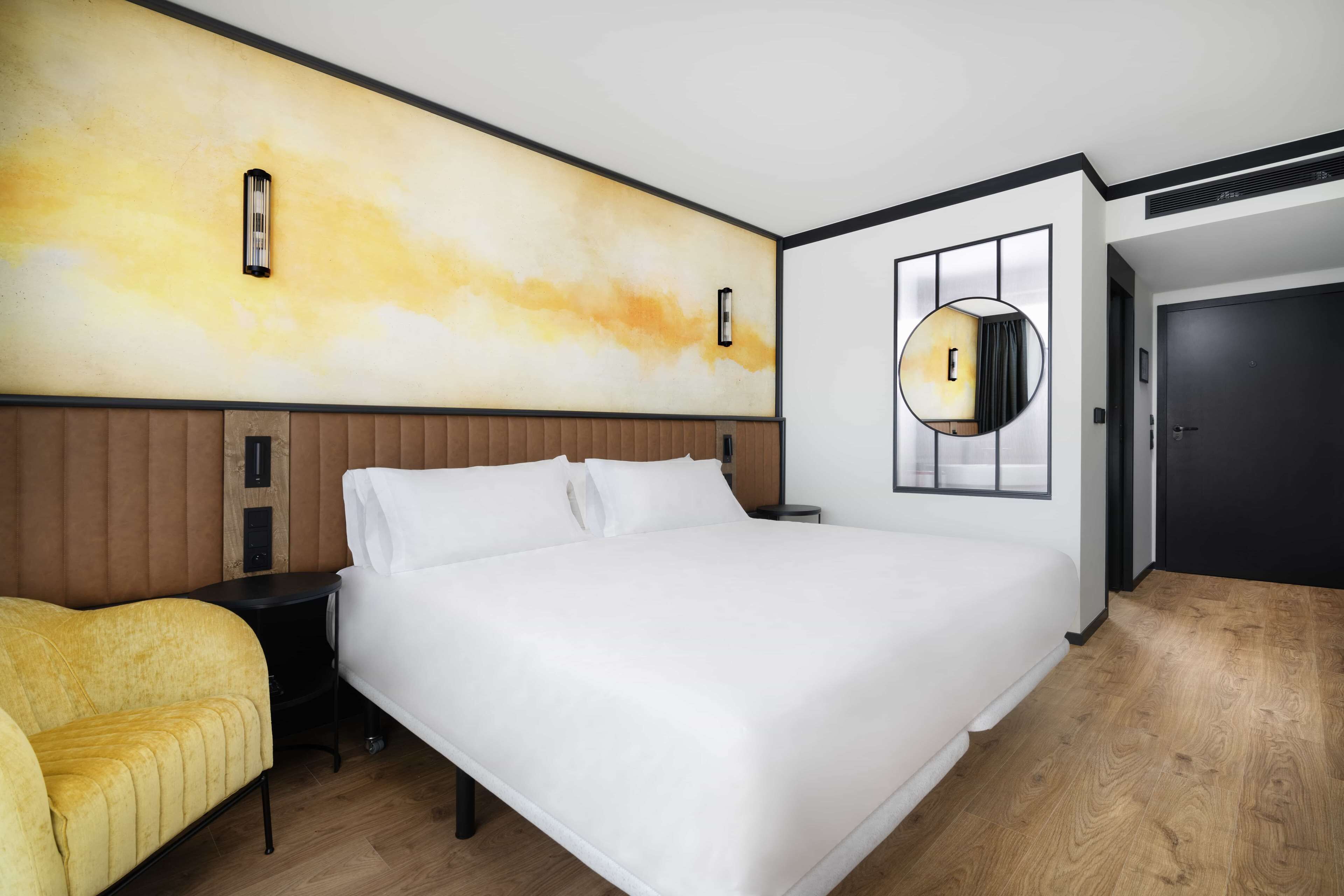 Une chambre d’hôtel avec un grand lit, une table de chevet, un fauteuil jaune, un miroir fixé au mur et une porte.