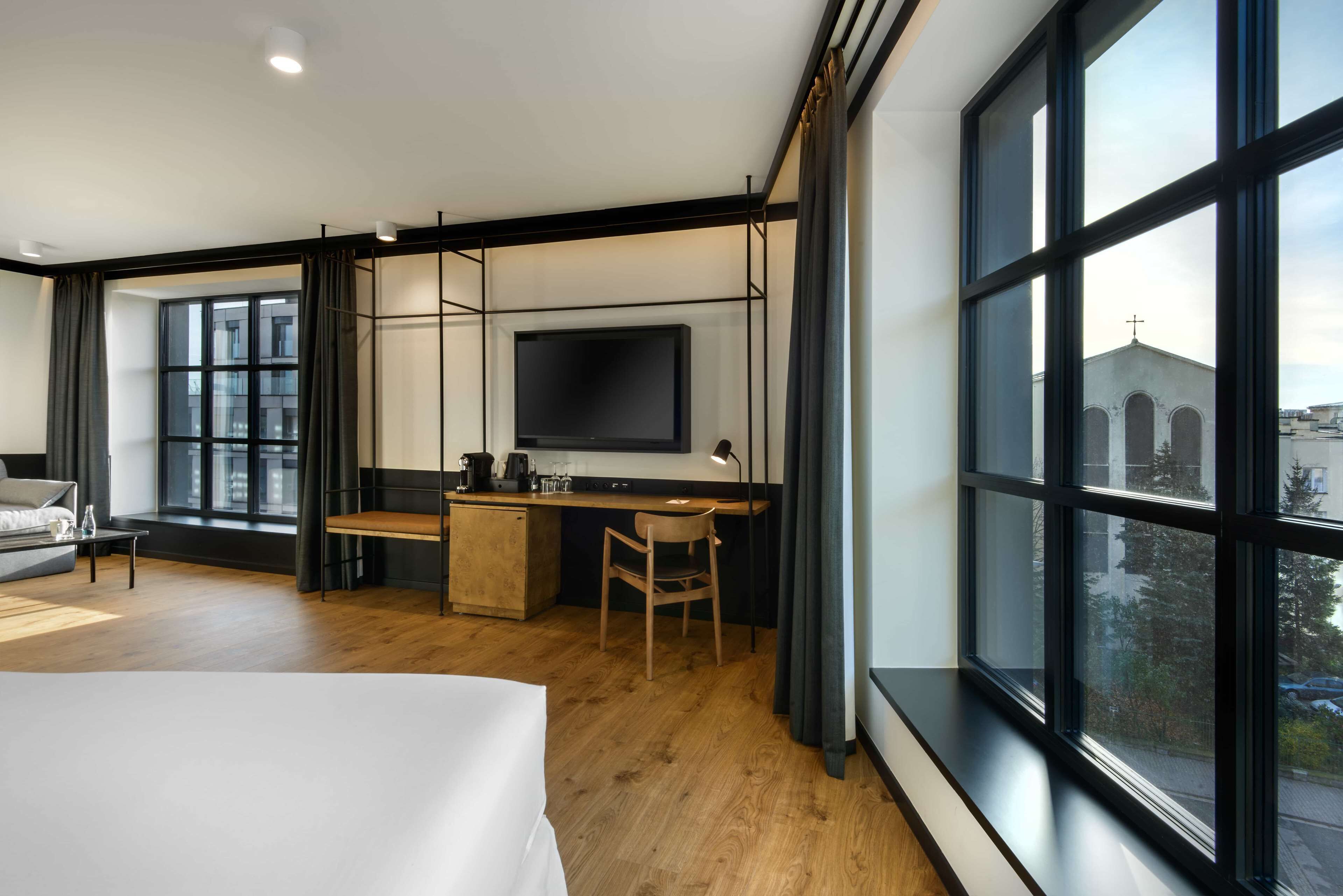 Ein modernes Hotelzimmer mit einem großen Bett, einem Schreibtisch mit einem Flachbildfernseher, einem Holztisch, einem Sessel und großen Fenstern mit Blick auf die Stadt.