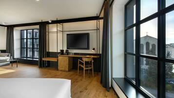 A modern hotel room with a large bed, a desk with a flat-screen TV, a wooden desk, a chair, and large windows offering a view of the city.
