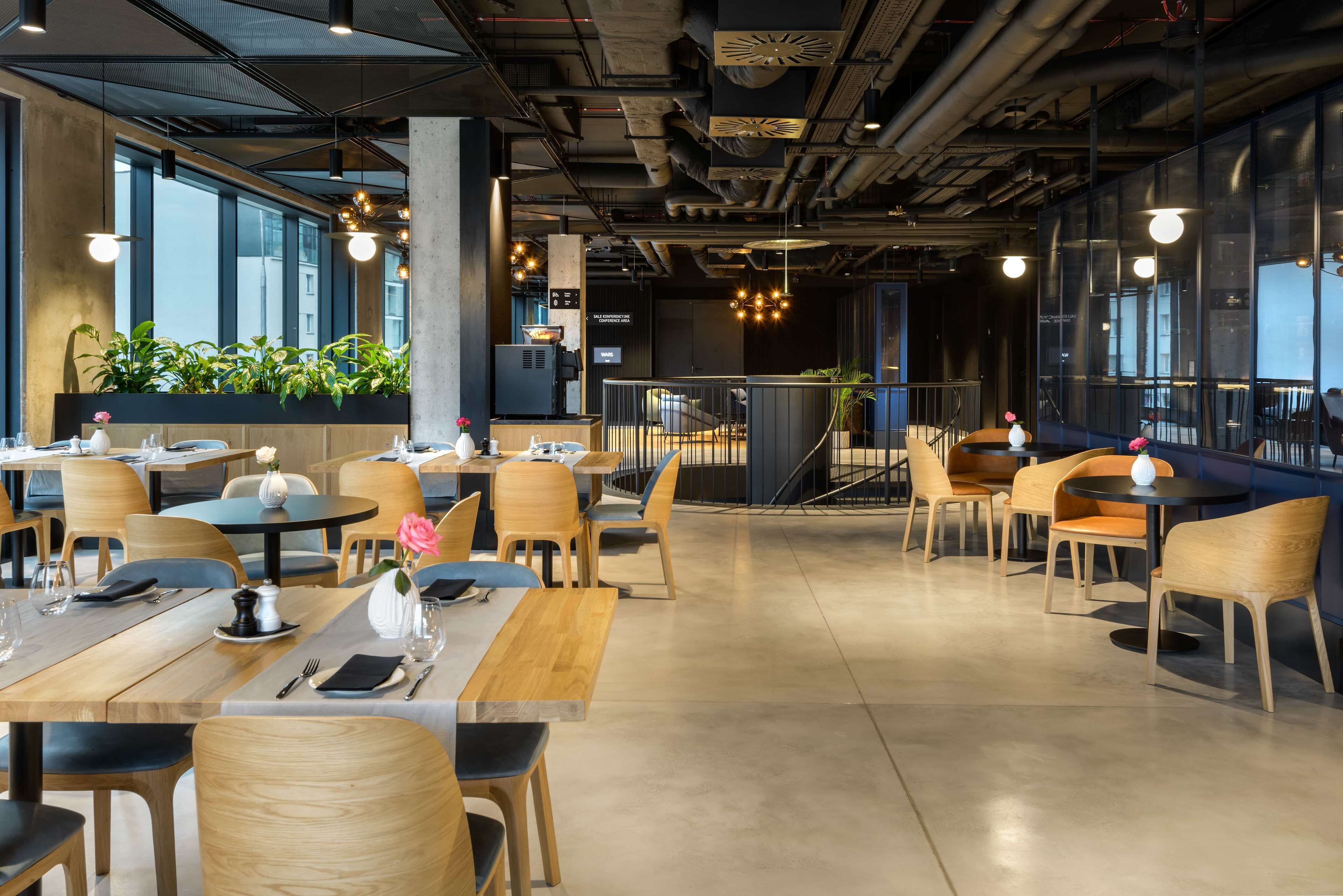 A modern restaurant with wooden tables and chairs, large windows, and indoor plants.