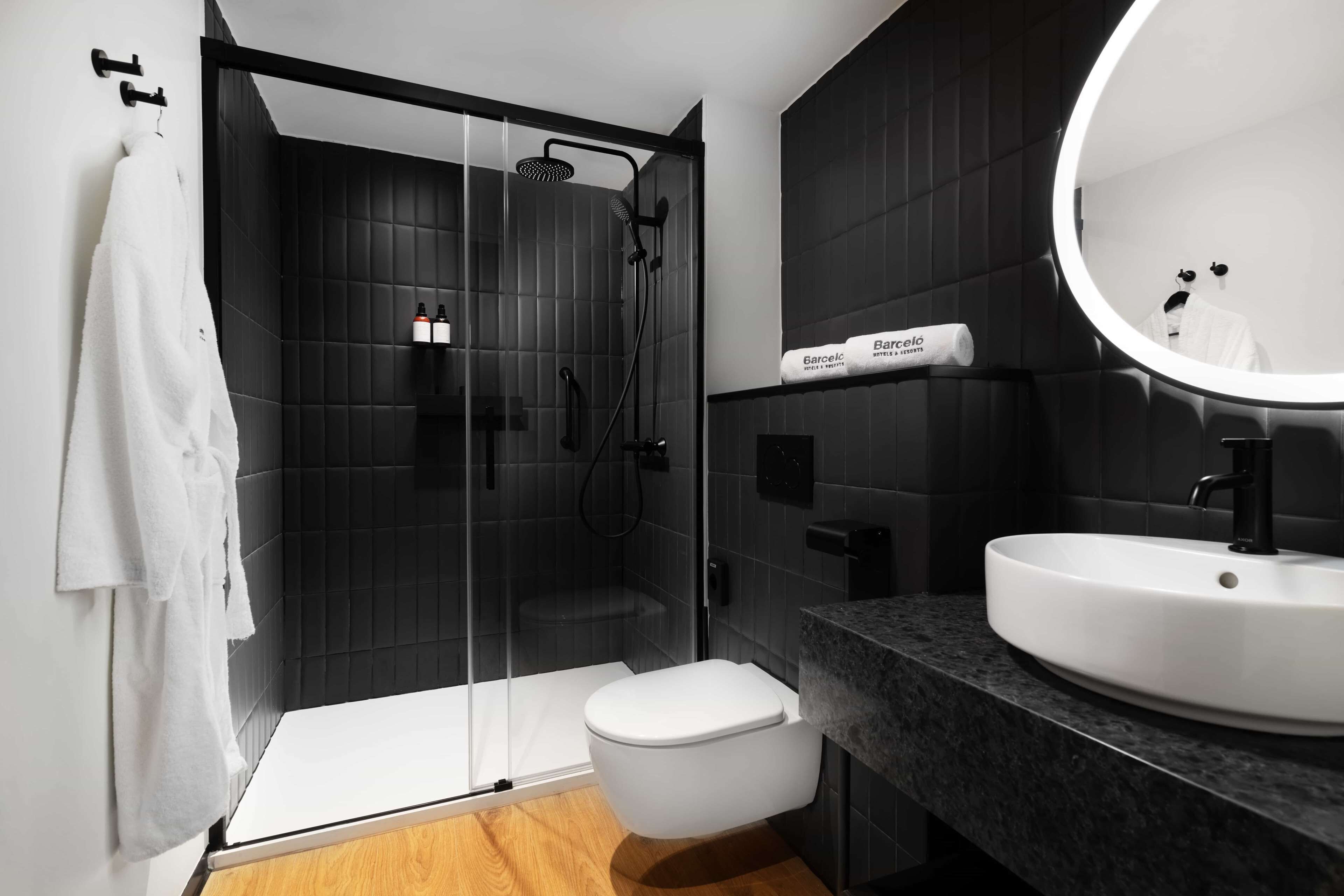 Un baño moderno con ducha envuelta en vidrio, inodoro blanco, espejo redondo y encimera negra.
