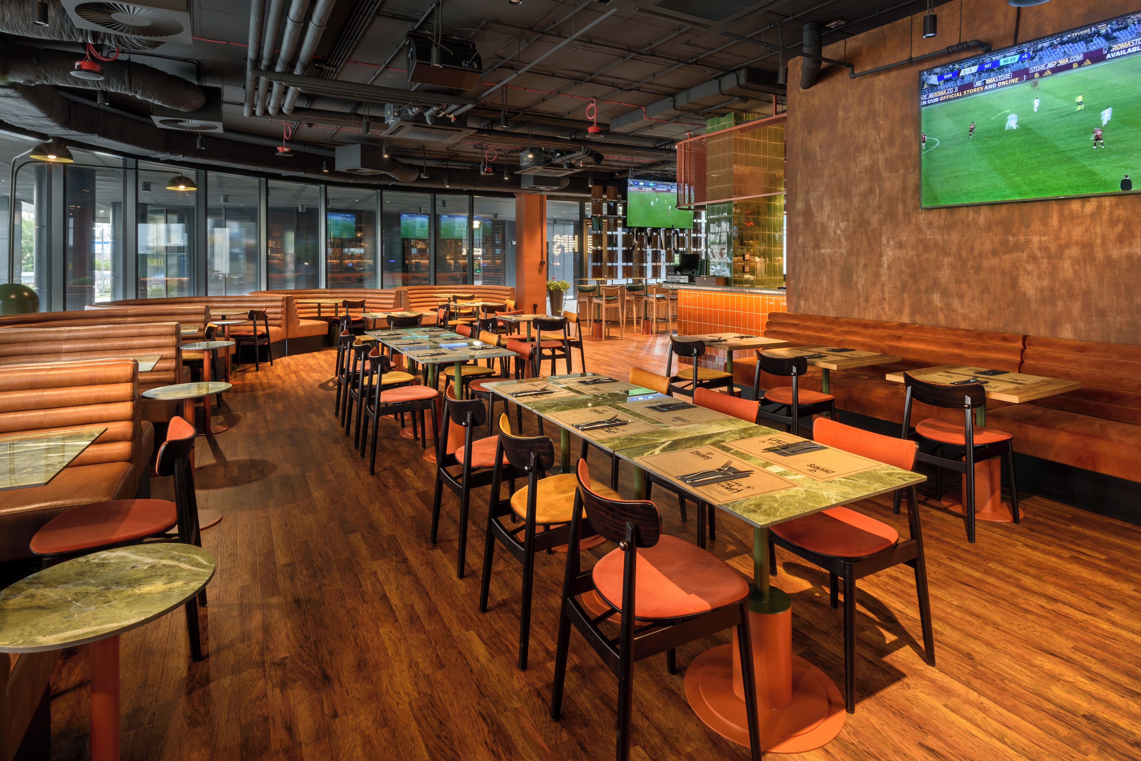 Un restaurant avec des banquettes, des tables et des chaises, et un grand écran de télévision diffusant un match de football.