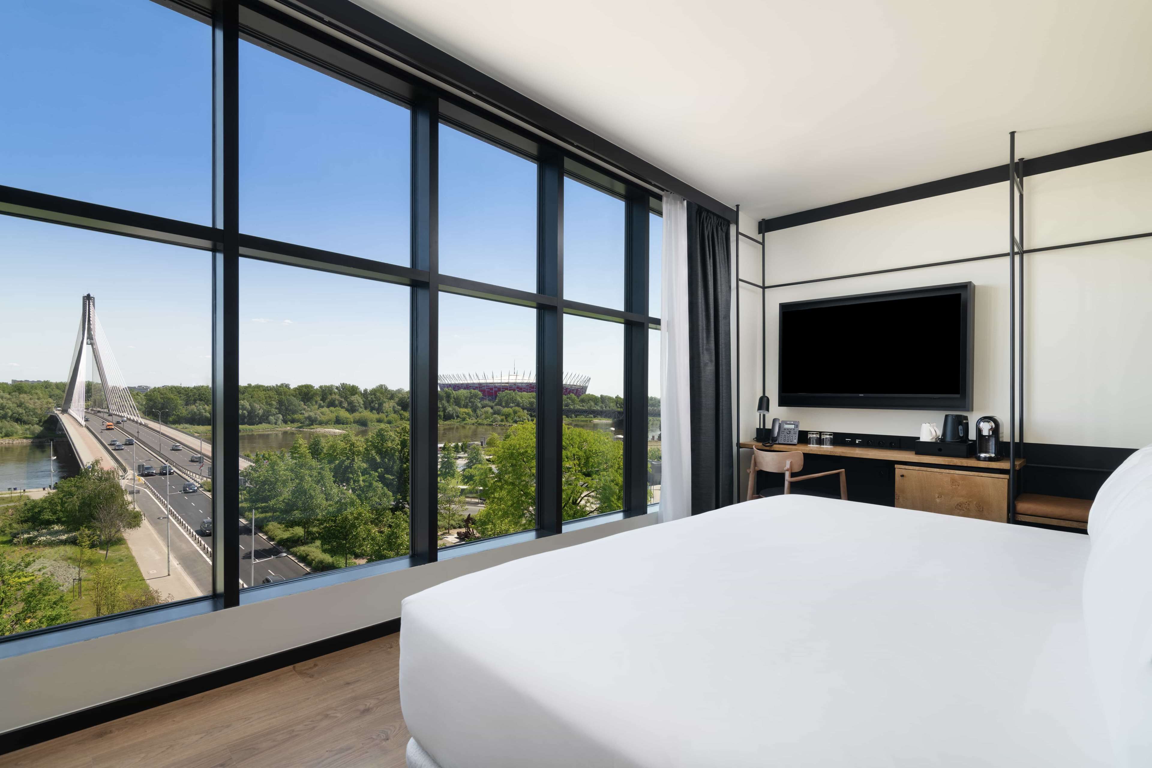 A modern hotel room with a large bed, a TV, a desk, and a view of a bridge and greenery.