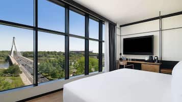 A modern hotel room with a large bed, a TV, a desk, and a view of a bridge and greenery.