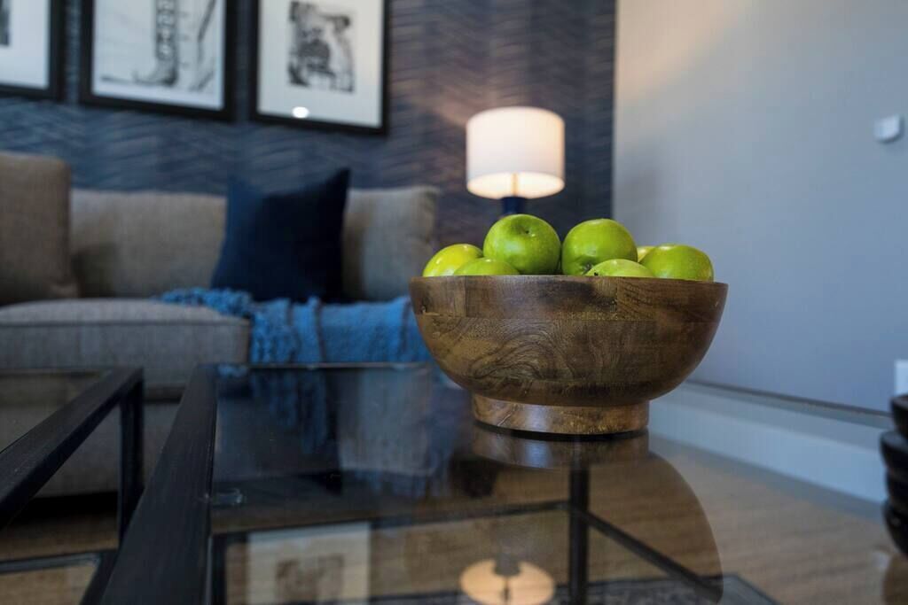 A wooden bowl with green apples on a glass table in a living room with a sofa and a lamp in the background.