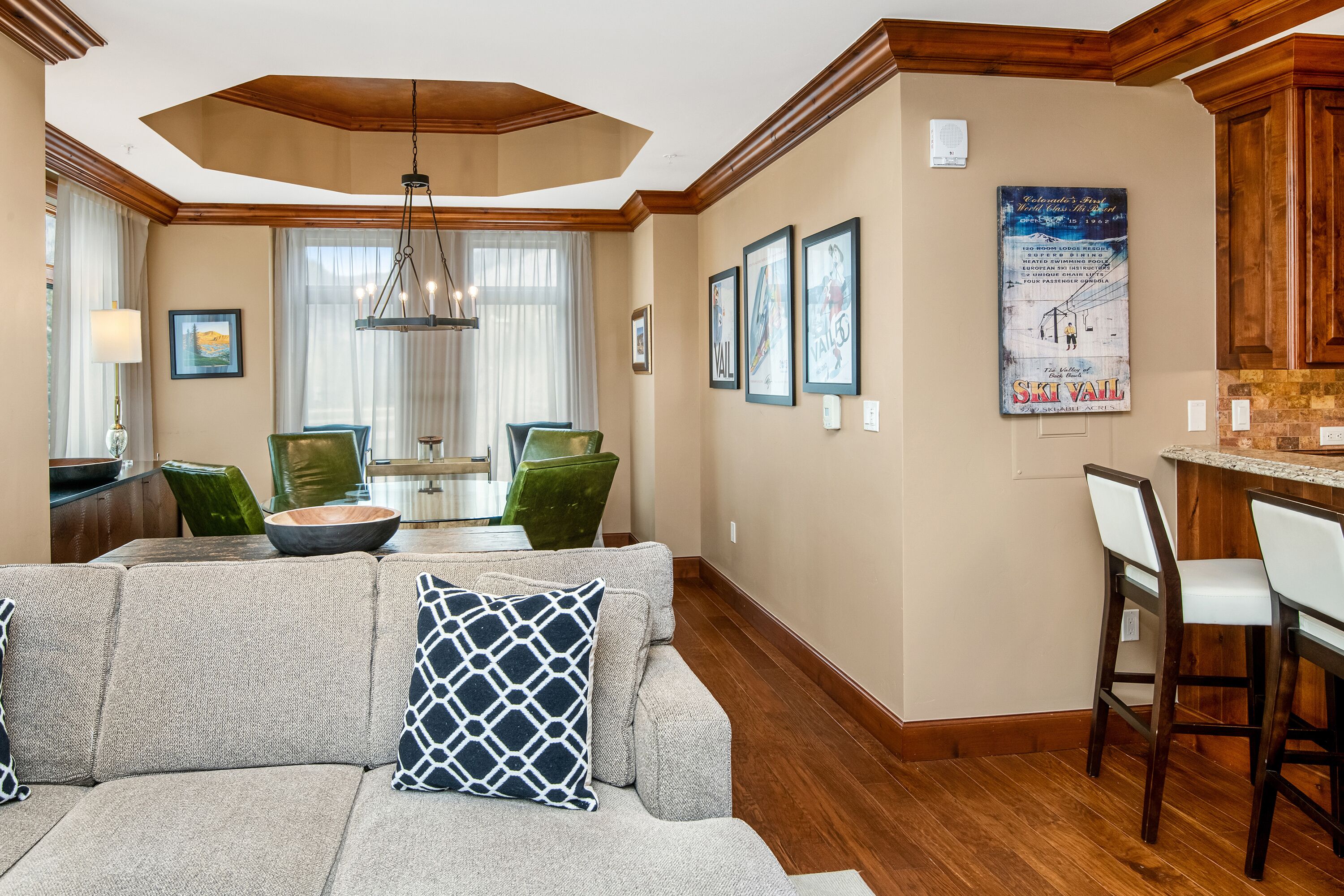 A modern kitchen with wooden cabinets, a dining area with green chairs, and a living room with a gray sofa and patterned pillows.