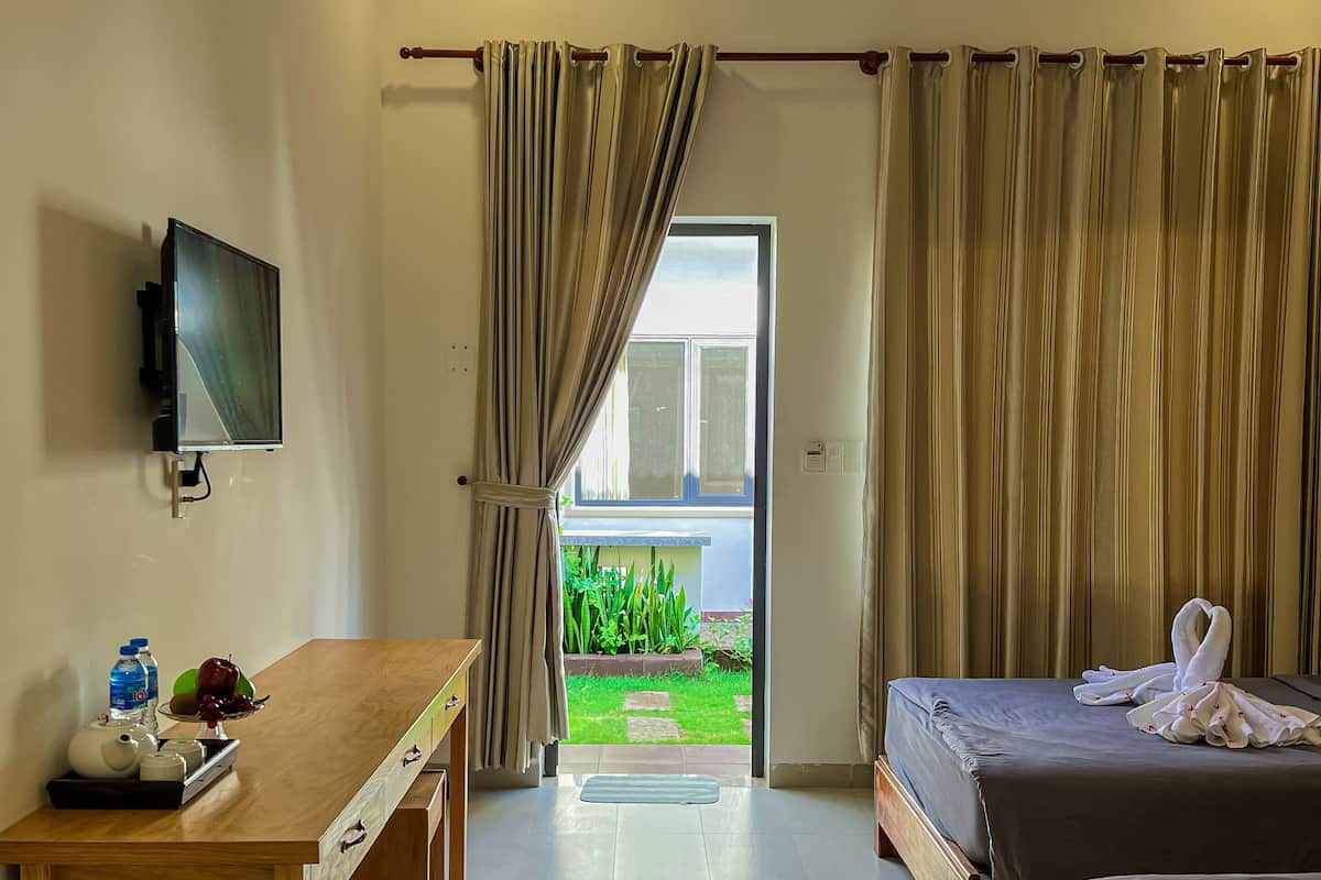 A hotel room with a bed, a desk with a TV, and a view of a garden through a door.