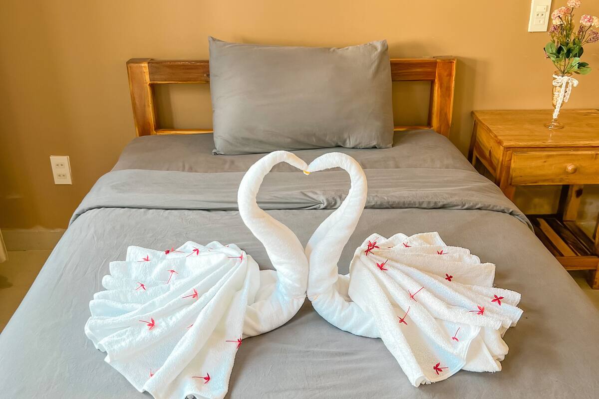 A bed with a grey bedspread and a pillow. There are two towels arranged in the shape of swans on the bed.
