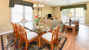 A dining area with a glass table and wicker chairs, a living room with a sofa and a TV, and a patterned rug on the floor. - 5 BR Beach House in Surfside Beach with Private Pool! C-Batical (Surfside Beach)