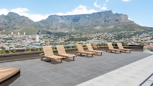 A rooftop terrace with wooden lounge chairs and a cityscape view of mountains and buildings. - Designer Apartment at the Peak of Bo-Kaap | Pool (Cape Town)