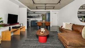 A modern living room with a wooden bench, a red stool, a brown leather couch, and a TV mounted on the wall. - Designer Apartment at the Peak of Bo-Kaap | Pool (Cape Town)