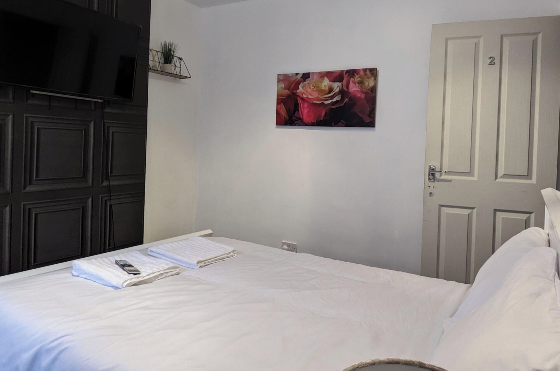 Een slaapkamer met een bed, een schilderij, een deur en een plank met een plant.