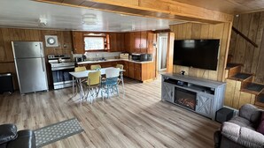 A spacious living area with a wooden floor, a dining table with chairs, a kitchen area with appliances, and a flat-screen TV mounted on the wall. - Cottage on Akers Pond (Errol)