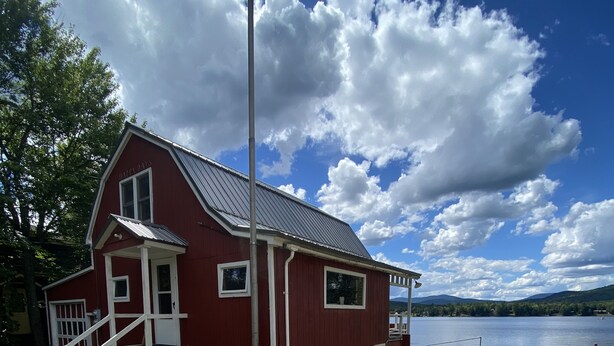 A red building with a porch and steps, near a body of water and mountains. - Cottage on Akers Pond (Errol)