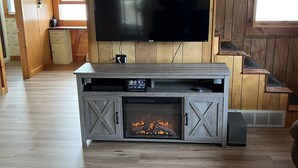 A living room with a large flat-screen TV, a built-in fireplace, wooden flooring, and a cozy seating area. - Cottage on Akers Pond (Errol)