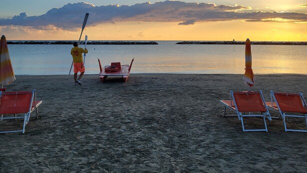 A beach with a person carrying a paddle, a row of beach chairs, and a row of umbrellas. - Vinsea Hotel (Bellaria-Igea Marina)