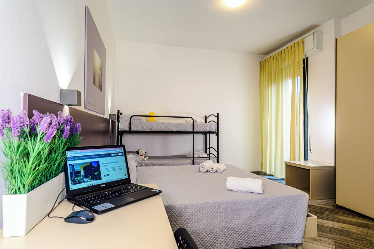 Una habitación de hotel con escritorio, ordenador portátil, literas, una mesita de noche pequeña y una planta en maceta.