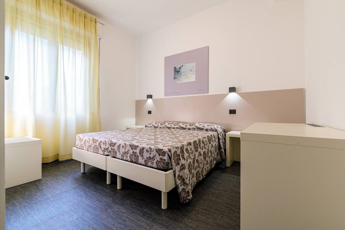 Una habitación de hotel con cama, mesitas de noche, escritorio y ventana con cortinas.