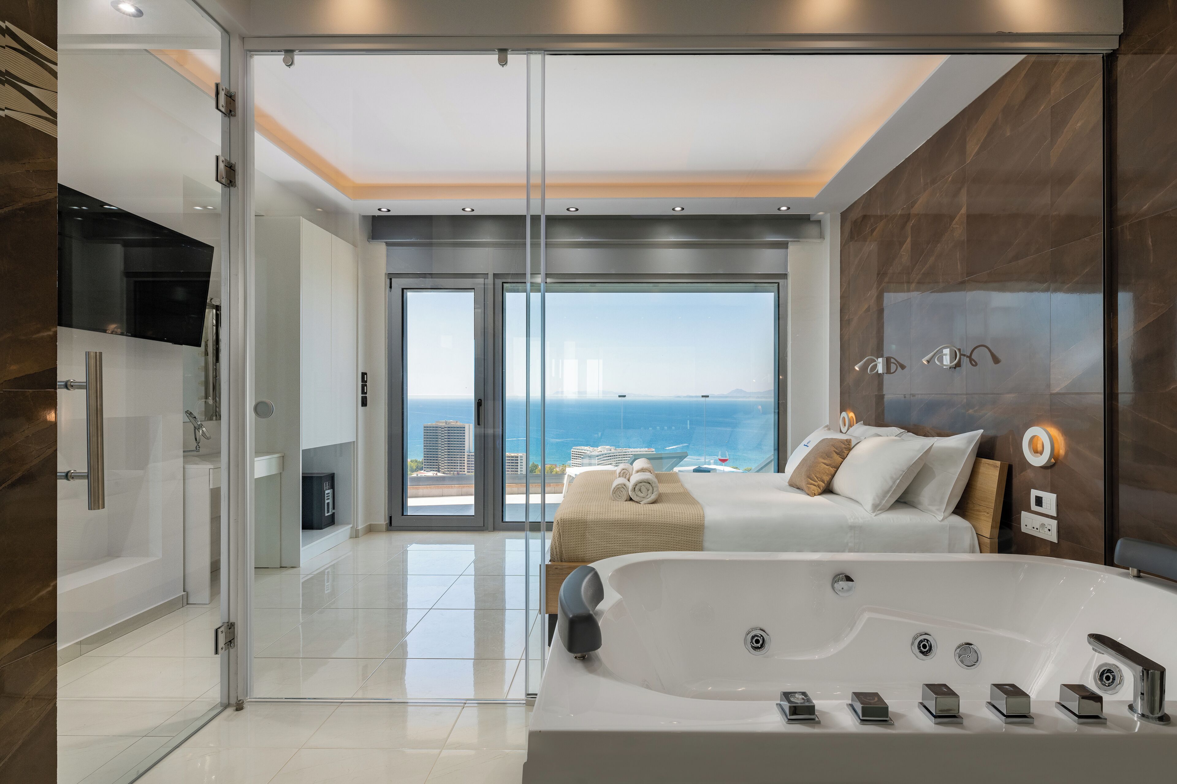Habitación de hotel moderna con una bañera grande, una cama con almohadas, una puerta de vidrio que da a un balcón y vistas al mar.