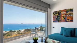 A modern living room with a large window, a blue sofa, a glass coffee table, and a painting on the wall. - Rhodes Skyline Suite With out Door Jacuzzi Sea View A2 (Rhodes)