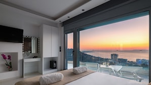 A modern bedroom with a large bed, a vanity with a mirror, and a view of the sunset over the water. - Rhodes Skyline Suite With out Door Jacuzzi Sea View A2 (Rhodes)