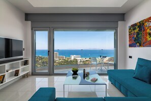 A modern living room with a large TV, a glass coffee table, a sofa, and a view of the sea.