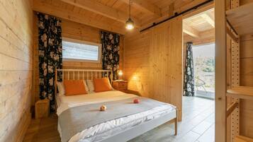 A wooden bedroom with a bed, a nightstand, a window with curtains, and a door leading outside.