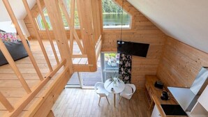A wooden cabin with a loft bedroom, a dining area with a table and chairs, and a large window overlooking a lake. - Scandinavian holiday home right by the lake (Klepnica)