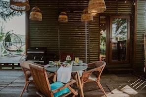 Eine Holzhütte mit einem runden Tisch, Korbstühlen, einem Grill und hängenden Lampen aus Korb.