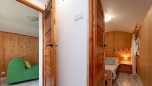 Una habitación con puerta de madera, sofá verde, cama con mesita de noche y repisa con objetos decorativos.