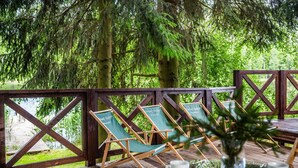 Terraza de madera con vista a un bosque, dos sillones verdes y una mesa con un florero y un menú.