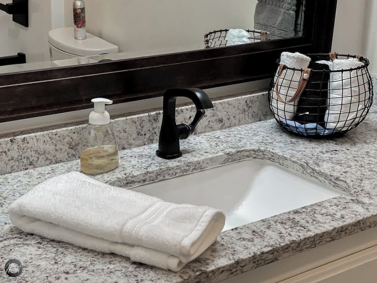 Une salle de bain avec un comptoir en granit, un lavabo blanc, un robinet noir, un distributeur de savon, une serviette et un panier en fil métallique contenant une autre serviette.