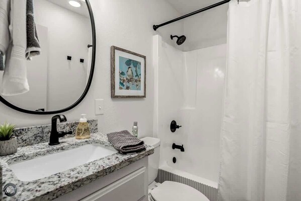 A bathroom with a granite countertop sink, a black faucet, a framed picture on the wall, a shower with a black curtain, and a white toilet.