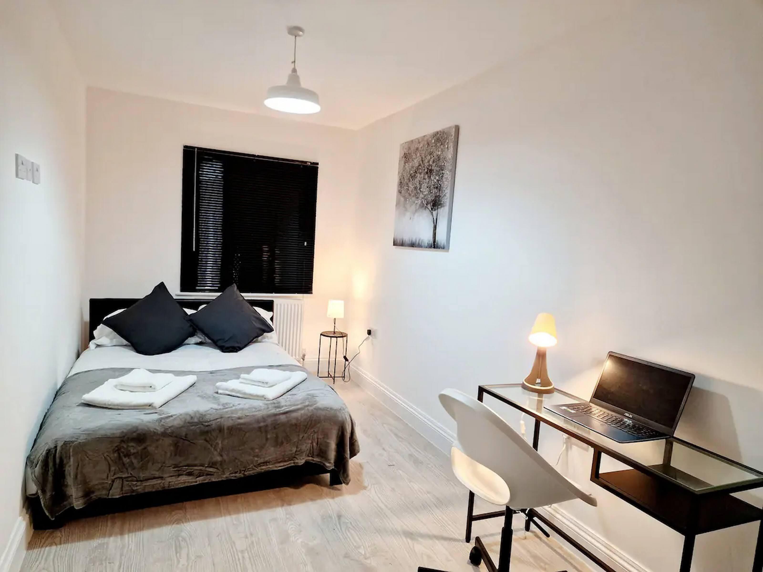 A bedroom with a bed, a desk with a laptop, a chair, a window with blinds, a wall-mounted lamp, and a painting.