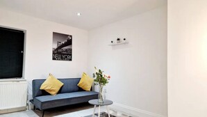 A modern living room with a blue sofa, two yellow cushions, a small round table with a vase, a black and white rug, and a cityscape poster on the wall. - Deluxe 2 Bed Apartment in Uxbridge (Uxbridge)