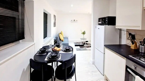A modern kitchen with white cabinets, a black countertop, and a microwave. There is a dining area with a black table and chairs, and a small plant on a side table. - Deluxe 2 Bed Apartment in Uxbridge (Uxbridge)