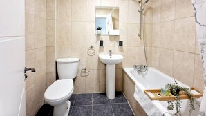 A compact bathroom with a toilet, sink, and bathtub, featuring beige tiles and a small mirror above the sink. - Deluxe 2 Bed Apartment in Uxbridge (Uxbridge)