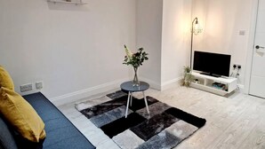 A modern living room with a flat-screen TV, a small round table with a vase of flowers, and a black-and-white rug. - Deluxe 2 Bed Apartment in Uxbridge (Uxbridge)