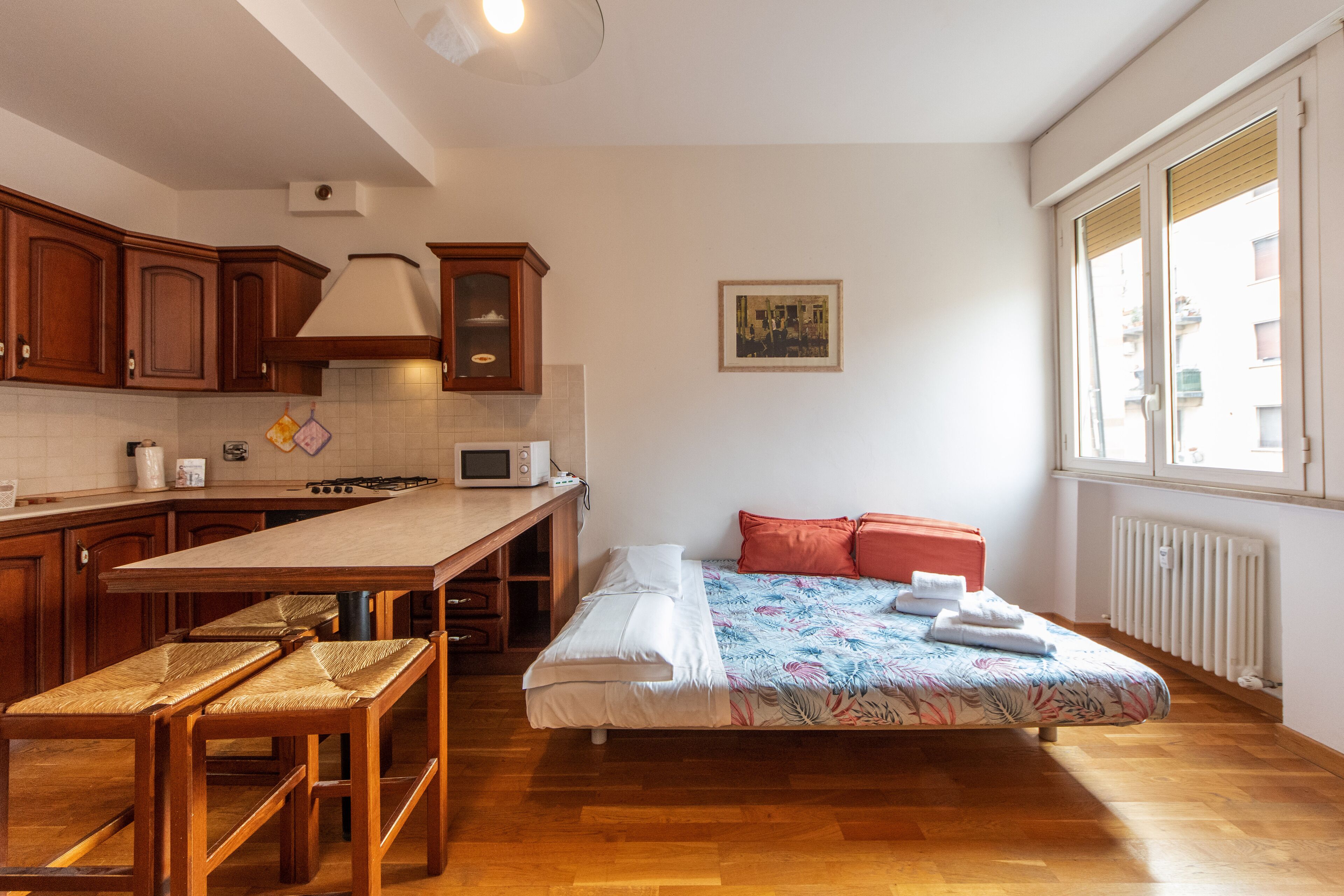 Habitació compacta amb cuineta, un llit amb edredó floral i una finestra amb vista a edificis.